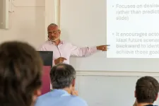 A Person standing behind a podium, holding a presentation and pointing at a board.